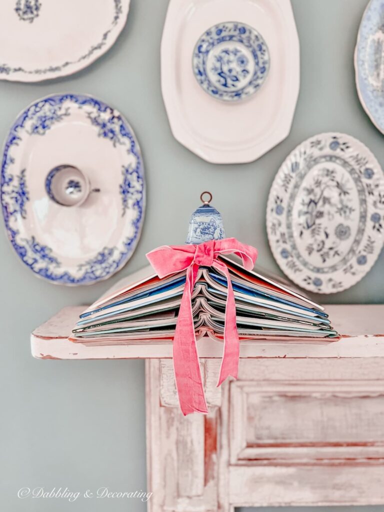 book Christmas tree made with six books and a pink bow topped with a vintage blue bell on a vintage mantel and blue and white platter wall gallery.