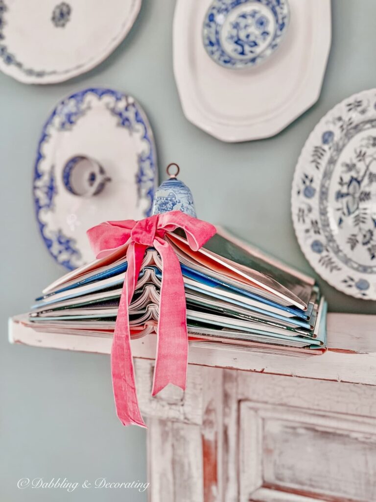 book Christmas tree made with six books and a pink bow topped with a vintage blue bell on a vintage mantel and blue and white platter wall gallery.