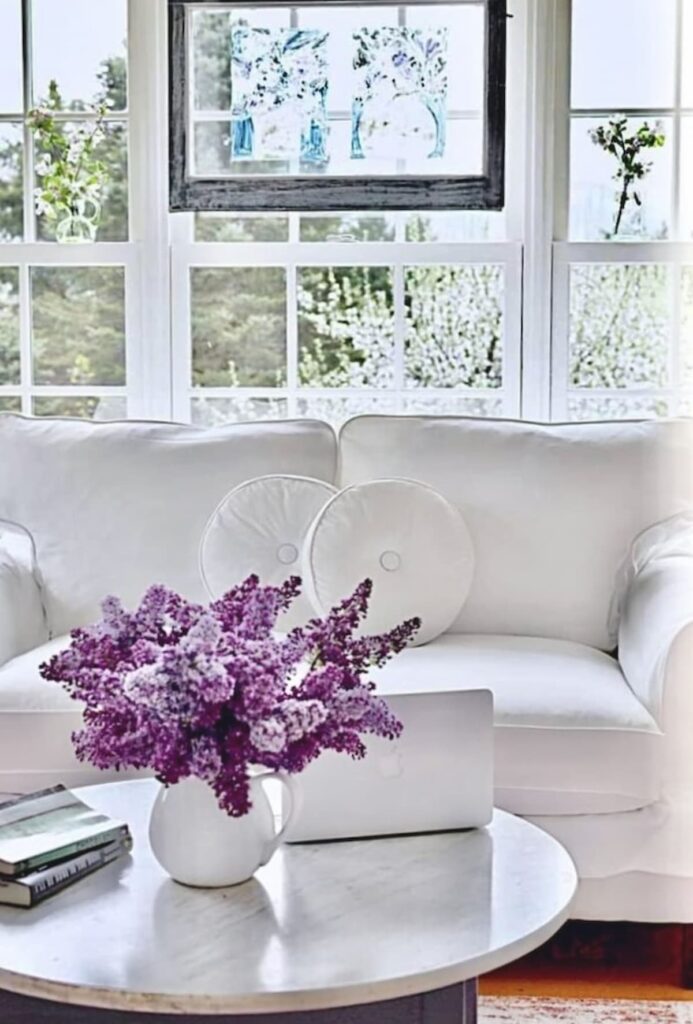 Sunroom Makeover Ideas with lilac flower bouquet and laptop open on marble coffee table with white slipcovered sofa and windows to spring.