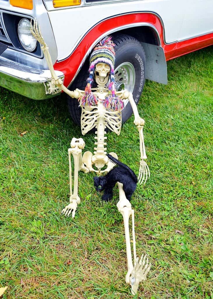 Halloween skeleton under truck tire. Skeleton on the ground in front of an old truck with a black cat and ski hat on for skeleton Halloween decorating ideas.