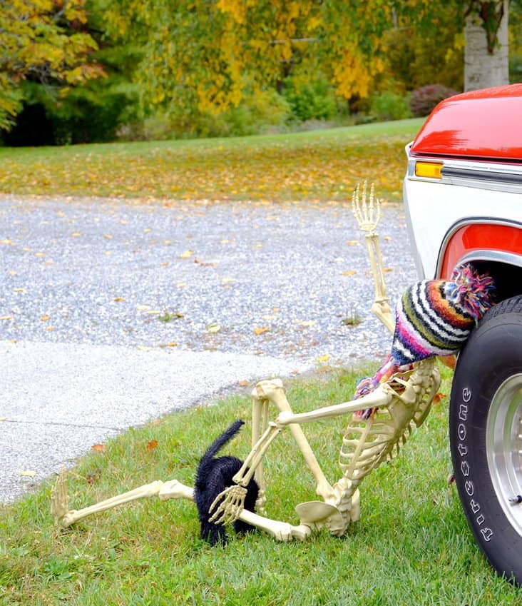 Halloween skeleton under vintage truck tire. Halloween decorating ideas with a skeleton on the ground with a black cat and hat on in front of an vintage red truck.