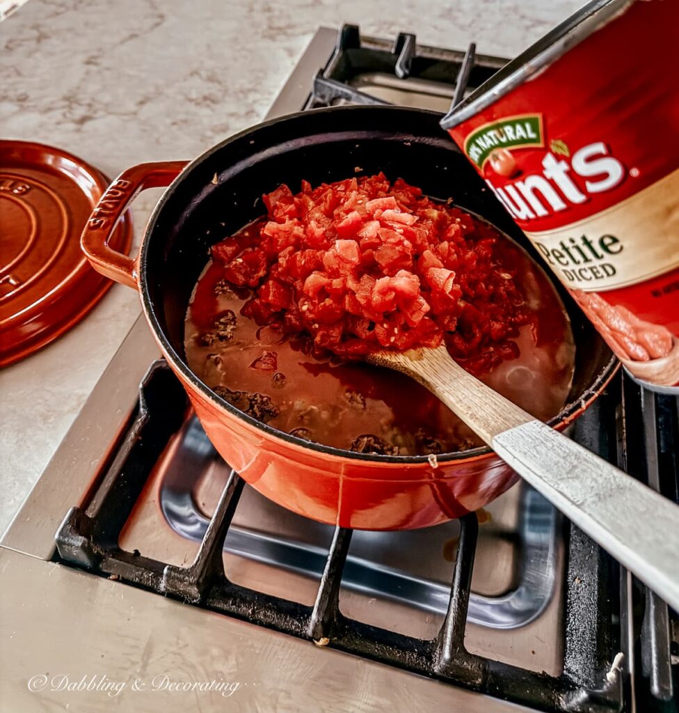 Adding diced tomatoes to a pasta bolognese sauce on stove with Hunt's can in hand and wooden spatula.
