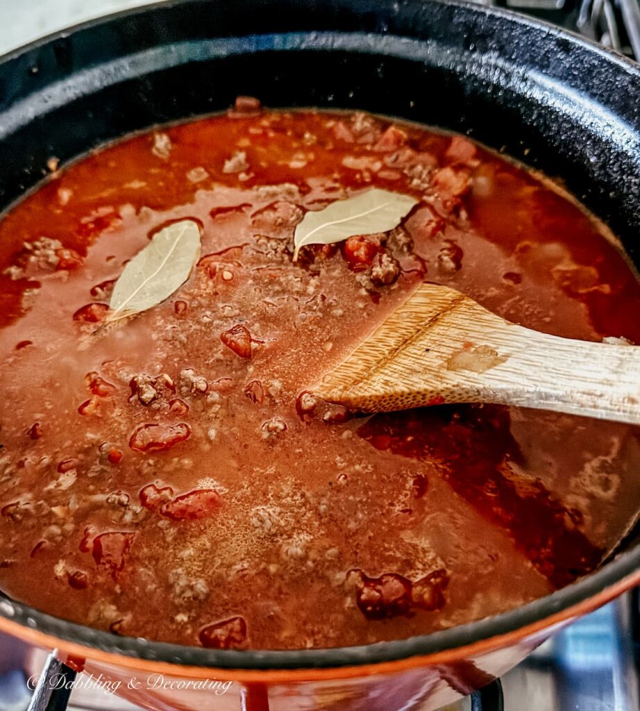 Authentic & Easy Pasta Bolognese Sauce Recipe Pasta Bolognese simmering on the stove with bay leaves and wooden spatula stirring the sauce.