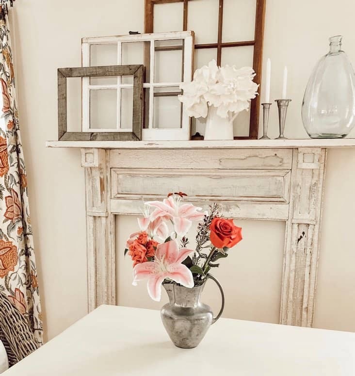 Vintage flower arrangement on white farmhouse table in dining room in front of vintage mantel on wall with layered old windows.