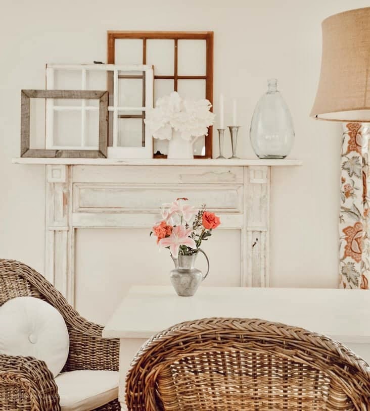 White vintage style dining room with wicker chairs, white farmhouse table, layered old windows and vintage flower arrangement after mantel install.