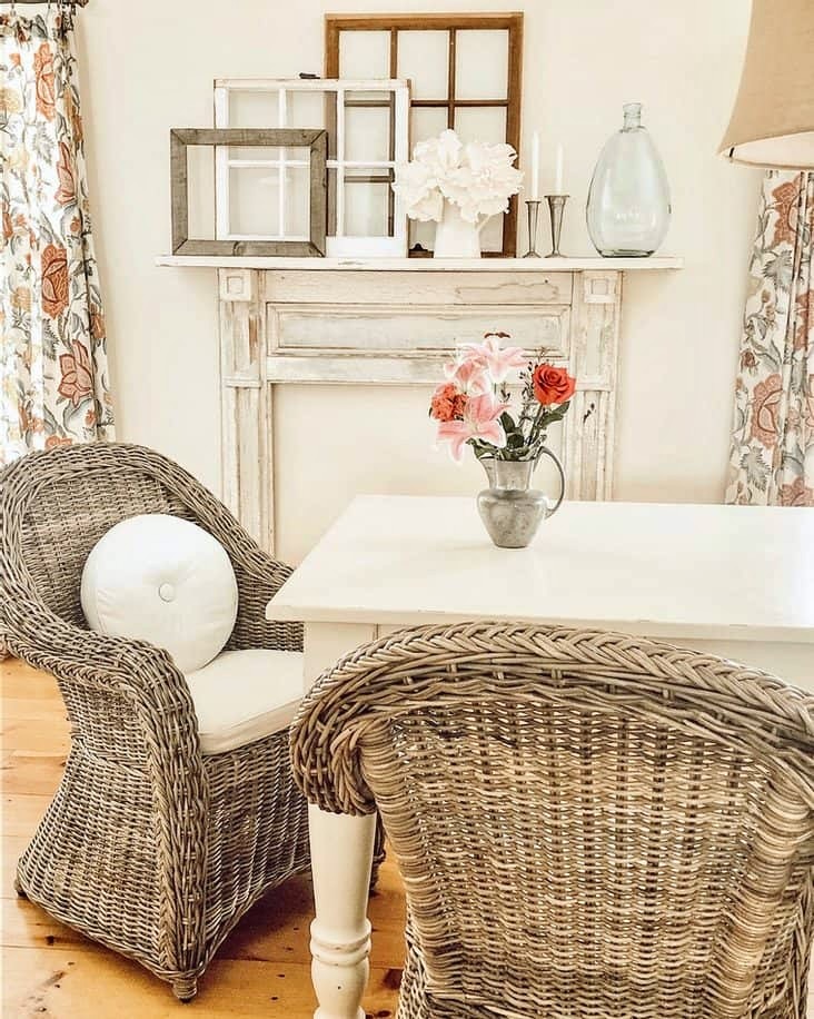 Vintage cottage style dining room with large white table, wicker chairs and vintage faux mantel install with layered old windows.