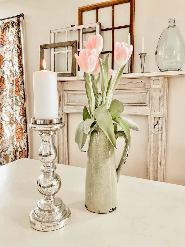Green pitcher with pink tulips, candle on table in front of vintage faux mantel install on wall.
