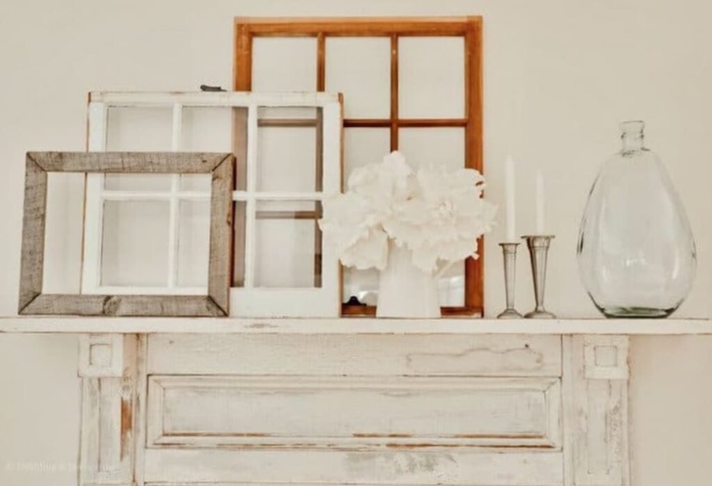 Three salvaged old windows layered on top of vintage white mantel with bottle and white flowers on dining room wall.