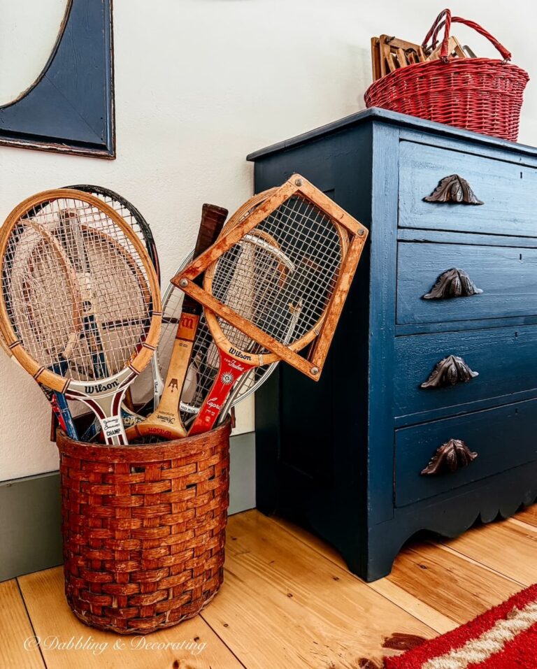 Antique basket with tennis racket decor next to a navy blue bedroom dresser on the floor.