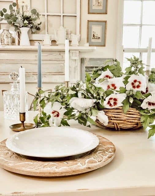 White flower centerpiece in basket on white styled farmhouse table with gold candlesticks, wooden charger and white plates.