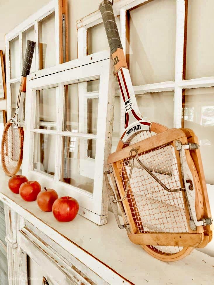 Vintage tennis rackets on mantel with salvaged windows and apples.