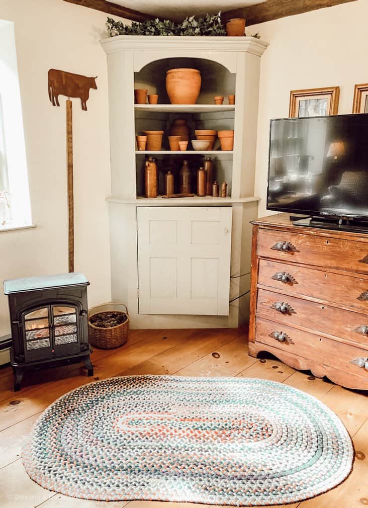 Antique corner hutch with terracotta pot collection, braided rug on wood floors, antique dresser with television in corner of living room.