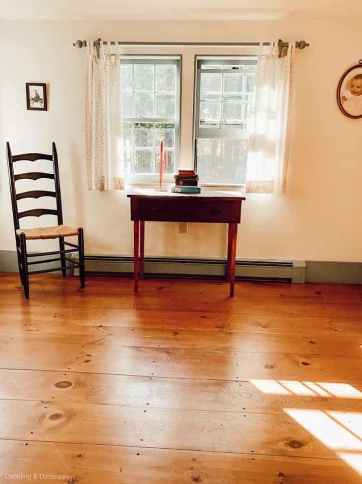 Empty guest bedroom with wood floors, chair and antique table with white curtains on windows before renovation.