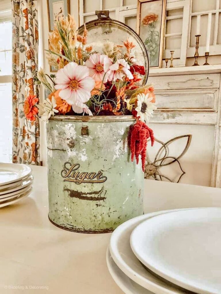 Flowers in a green antique sugar tin on white farmhouse dining room with faux mantel and white dishes on table.