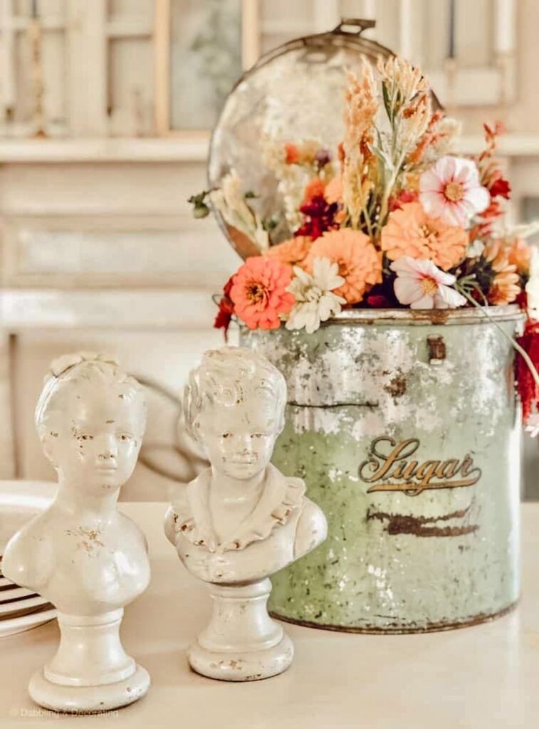 tin decor with old sugar tin with seasonal flowers and white busts on table for centerpiece.