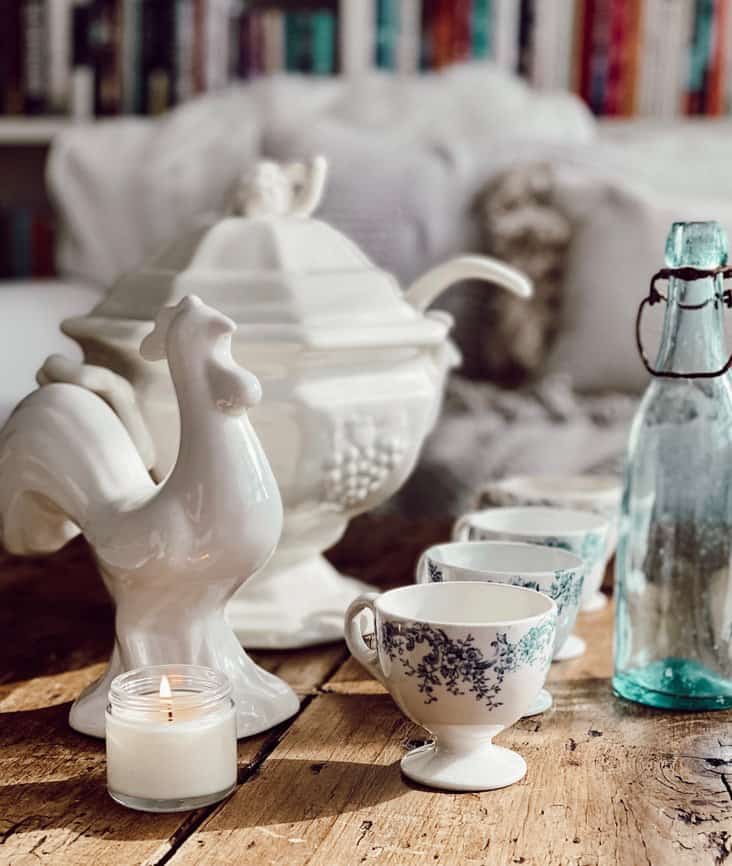 Antique finds on table, white soup tureen, white rooster, four teacups and blue bottle.