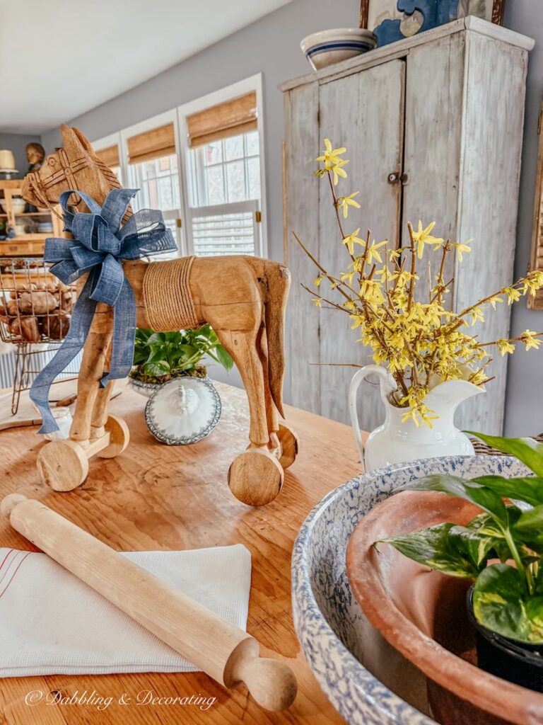 Antique wooden table with Forsythia Cut Flower Arrangement for spring next to a vintage wooden horse with a blue bow in a scullery-inspired kitchen space.