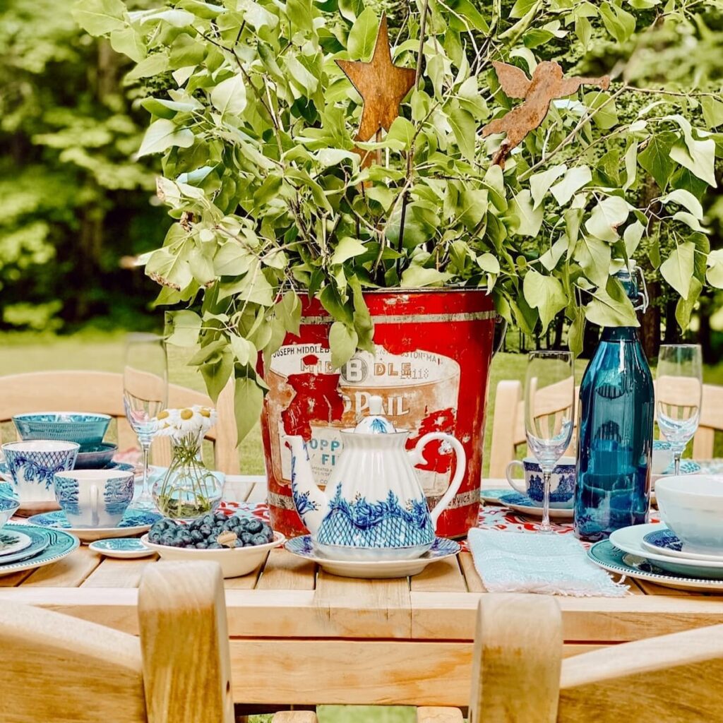 Patriotic centerpiece made with a chippy red galvanized bucket and green leaf branches paired with red, white, and blue place settings on an outdoor Patriotic table.