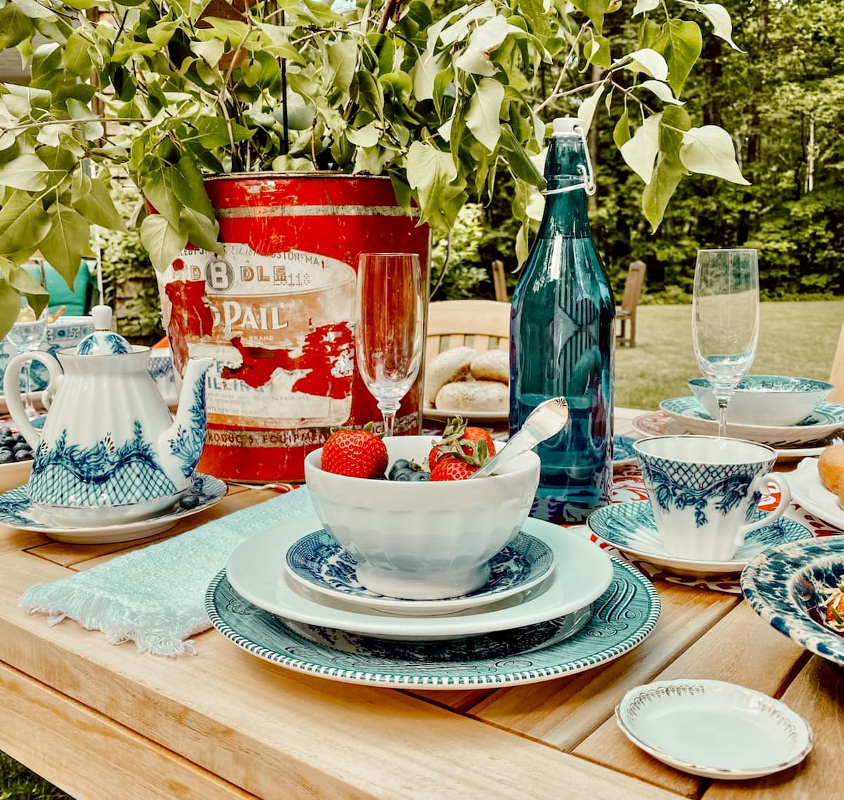 An Alfresco Summer Breakfast Table Red galvanize bucket with green branches on outdoor summer table with layered red, white, and blue place settings for breakfast.