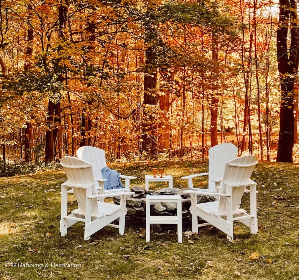DIY stone fire pit with white Adirondack chairs in backyard with beautiful fall sunset in the woods.