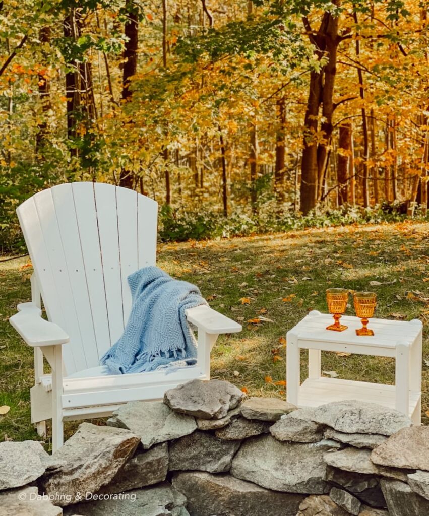 DIY stone fire pit in backyard with white Adirondack chair and table with blue blanket with fall sunset in the woods.