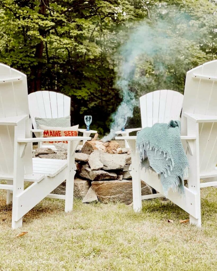 Stone fire pit in backyard with white Adirondack chairs and fire going.