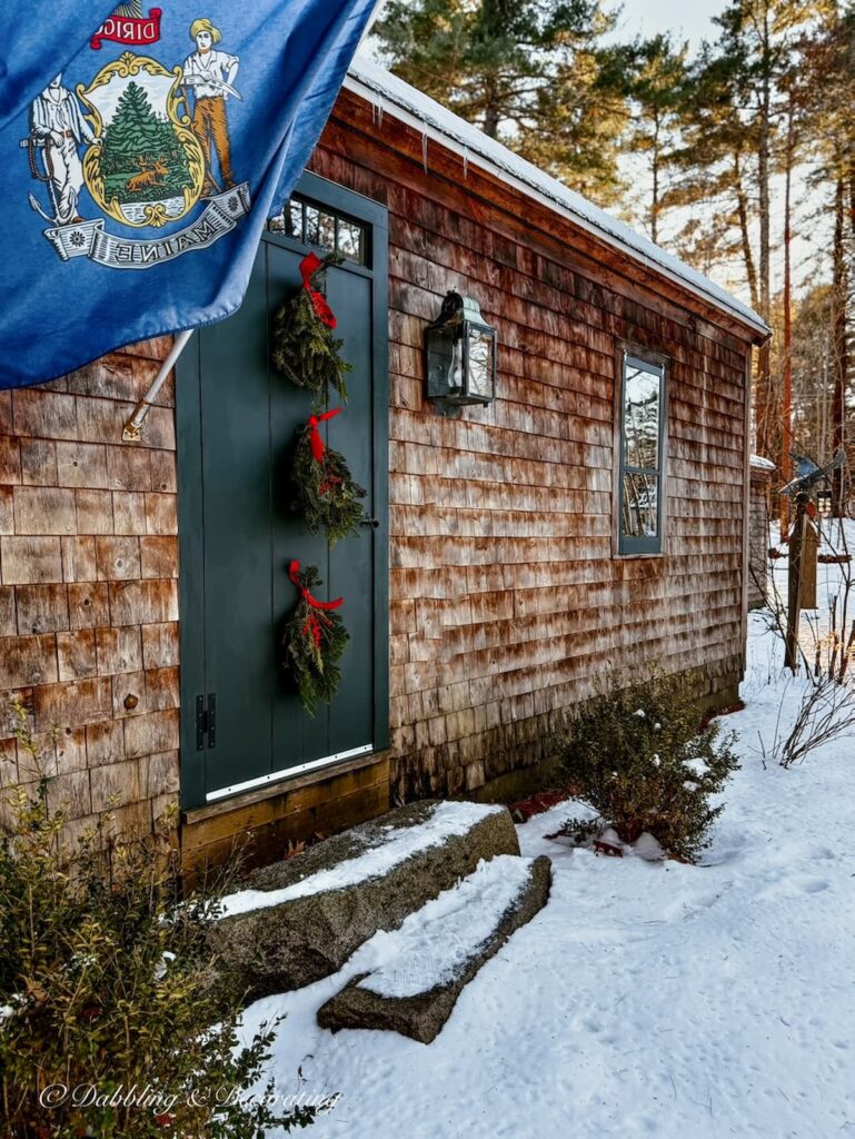 Three Christmas swags with red bows hanging on the front door of a cedar shakes home in Maine.