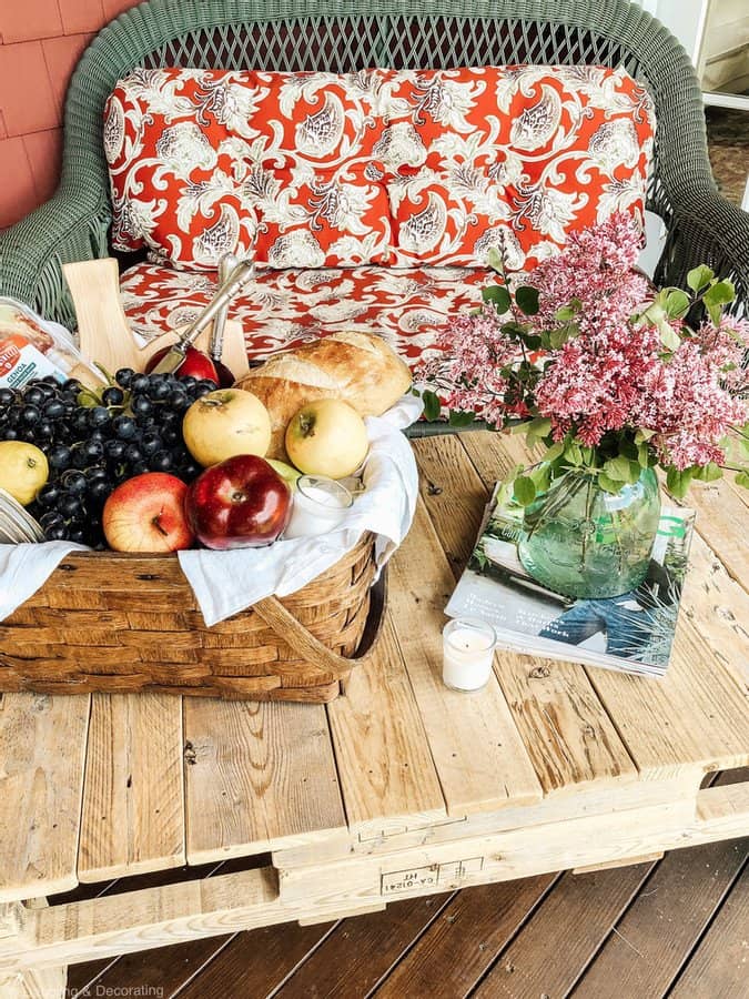 Picnic set up on DIY pallet coffee table with flowers on wicker couch with red cushions on porch.