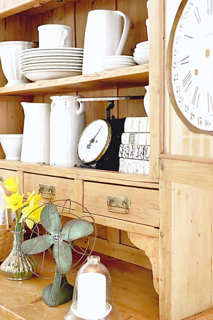 White pottery in wooden vintage hutch with vintage clock, scale, fan, candle and basket.