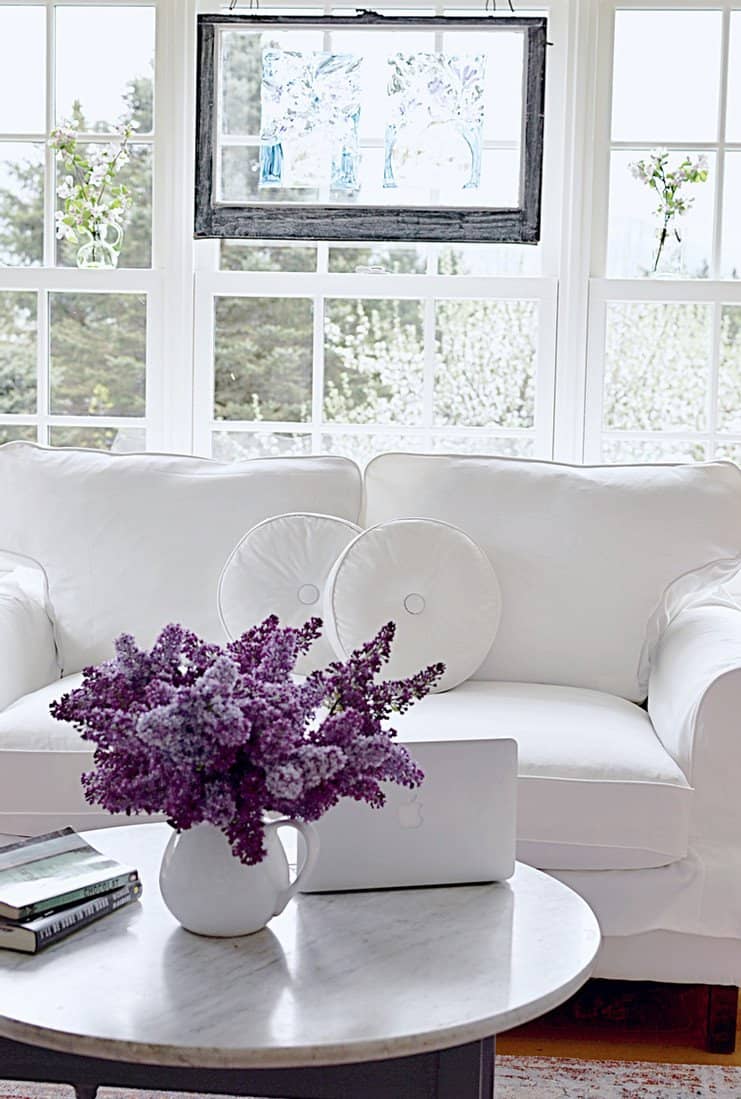 Vintage window hanging in window with vintage jars and white flowers with white loveseat and bouquet of lilacs.