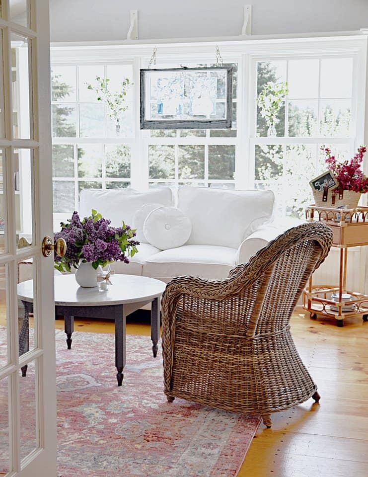 Sunroom makeover with white loveseat, vintage window hanging and a lot of flowers.