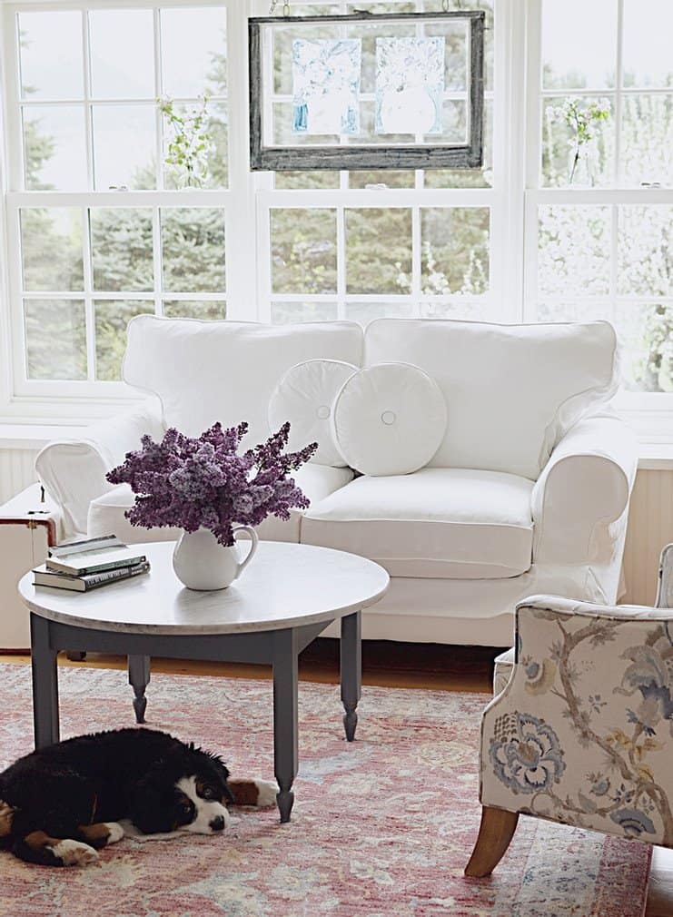 Vintage window hanging in window framed with white flowers in vintage jars, white loveseat, bouquet of lilacs and pink rug.