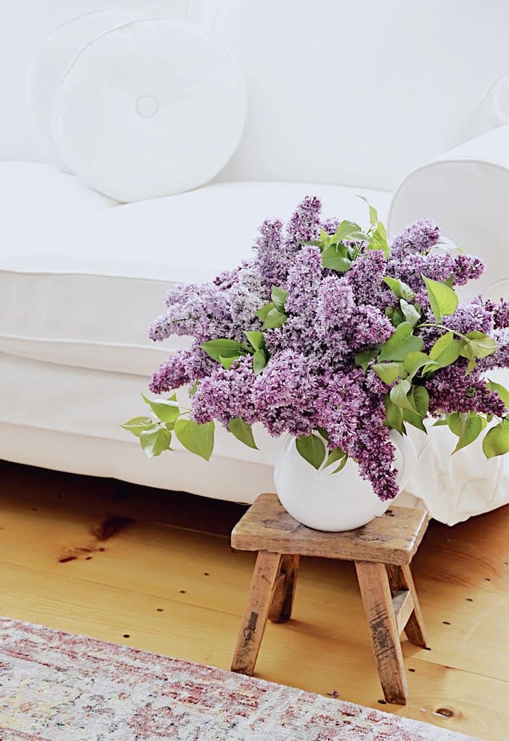 white loveseat with retro white pillows and a bouquet of lilac flowers on a vintage wooden stool.