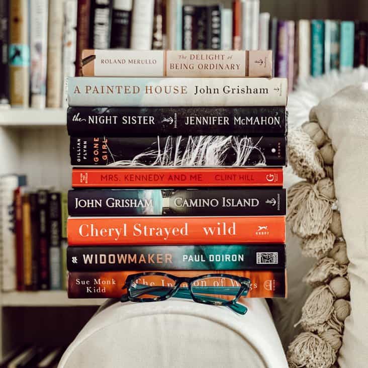 Stack of books on arm of couch with eye glasses in front of bookshelves.