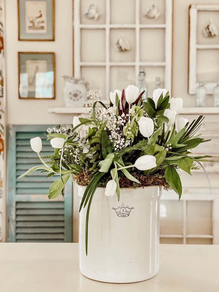 Vintage Crock White tulips and greenery spring floral arrangement on white table.