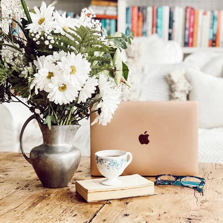 Home Apple laptop open with vintage cup of tea, book, eye glassware and pew pitcher of flowers on coffee table.