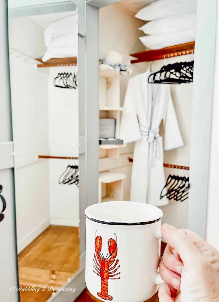 Small open closet with bathrobe and black velvet hangers and hand with lobster mug in bedroom.