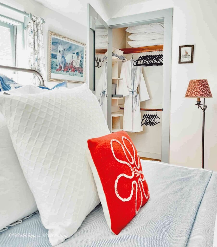 Red and white pillows on bed looking onto open closet with velvet black clothes hangers and bathrobe in small guest bedroom.
