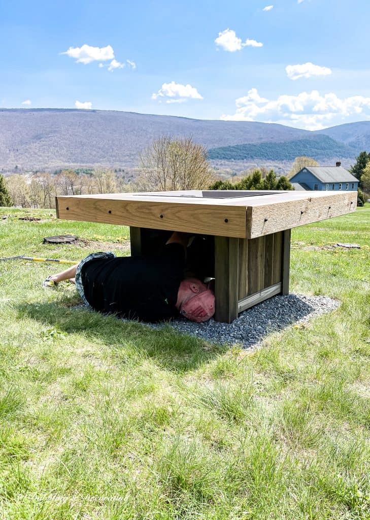 A wooden fire pit sitting in the grass with man install propane tank.