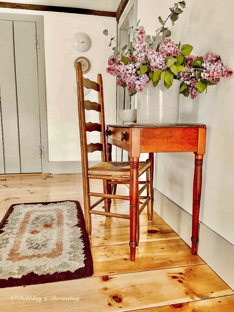 large lilac bouquet arrangement in antique crock on wooden antique table with chair and antique rug on wood flooring.