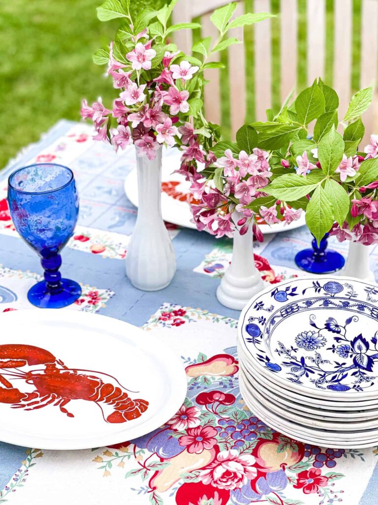 Outdoor summer tablescape with milk glass vases, lobster plates, vintage tablecloth and blue and white dishes.