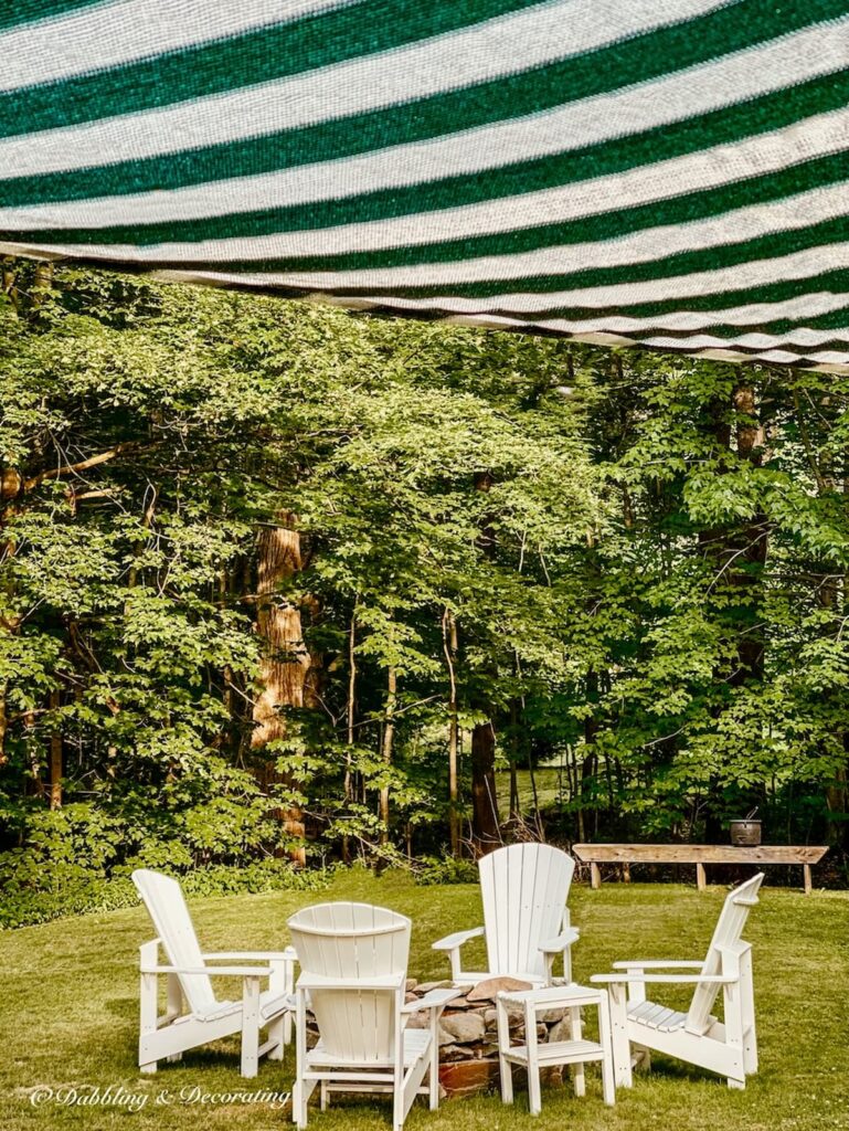 Backyard fire pit with white Adirondack chairs with green and white striped sun shade sail.