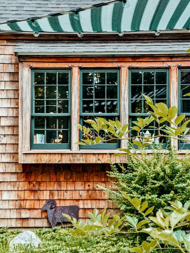 Cedar Shakes home's four windows trimmed in green with green and white striped sail shade with lights.