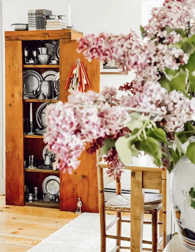 A vintage vase of lilac flowers next to an antique cabinet filled with pewter.