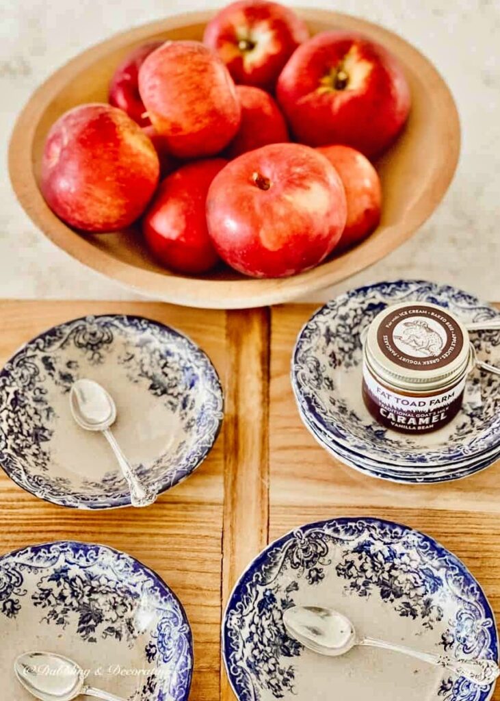 Wooden bowl of apples next to small blue and white vintage bowls on cheese board with silver spoons.