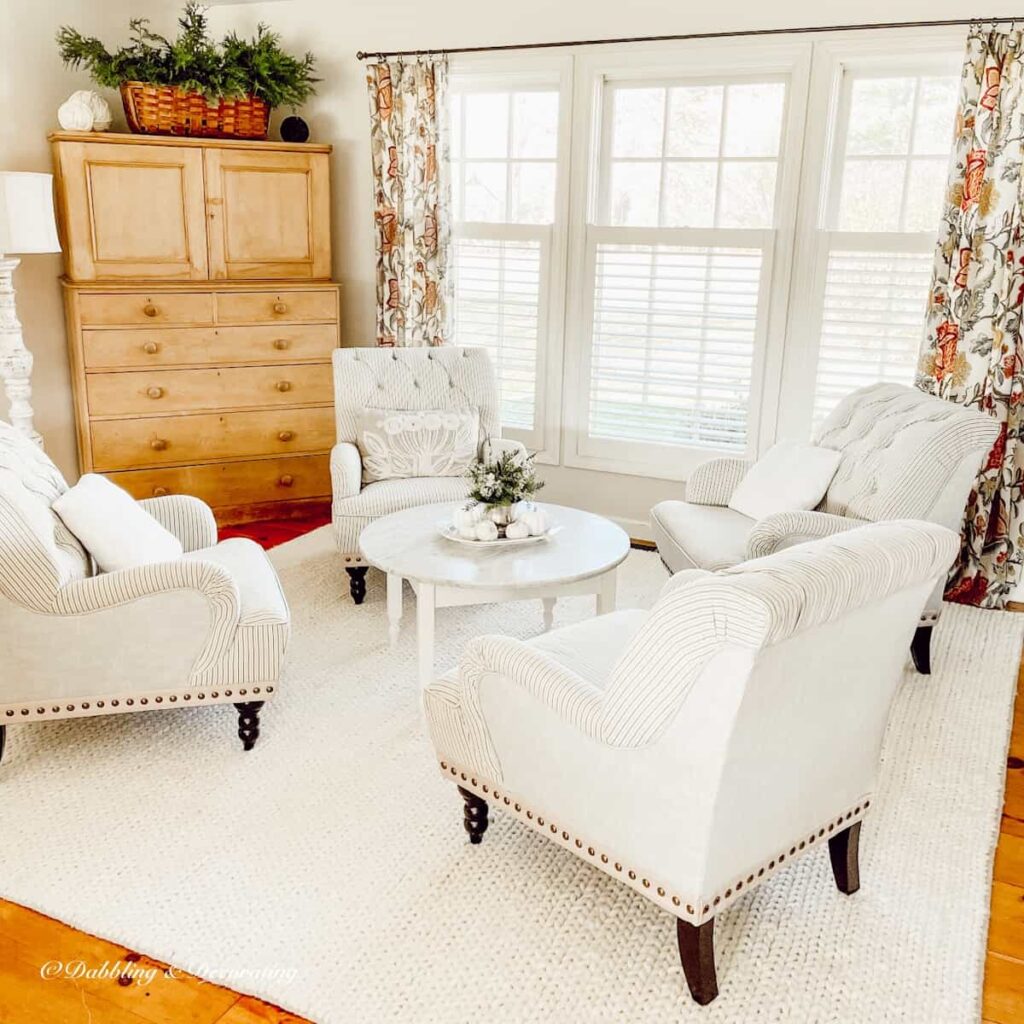 Sitting room with four white and green striped chairs on white rug, with marble coffee table and vintage pine hutch with colorful window curtains.