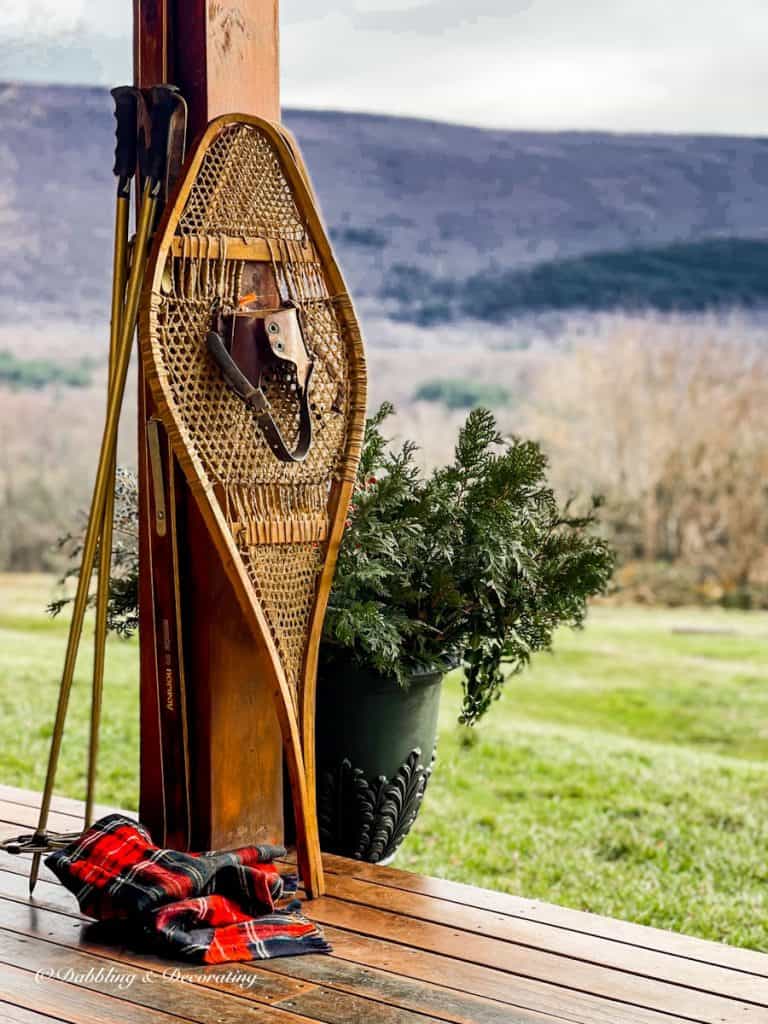 Vintage snowshoe on porch beam with red scarf on ground, ski poles, and evergreens in urn.