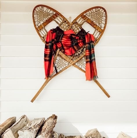 Wooden antique snowshoes on wall with plaid red scarf and firewood underneath on winter porch.