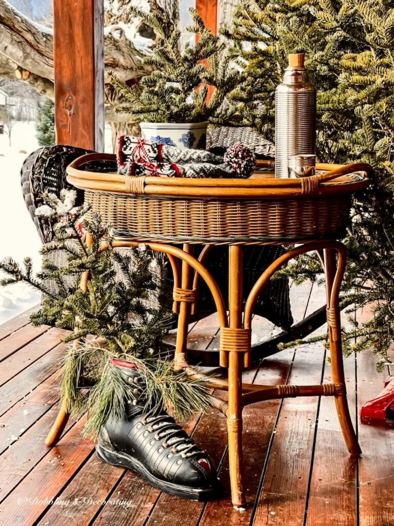 Outdoor porch wicker table with evergreens, vintage ski boot and old thermos and hat with snow in apres ski decor.