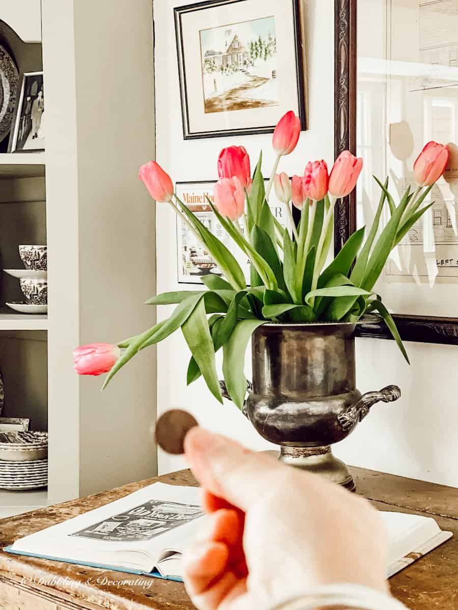 Hand holding up a copper penny in front of pink tulip bouquet on desk.